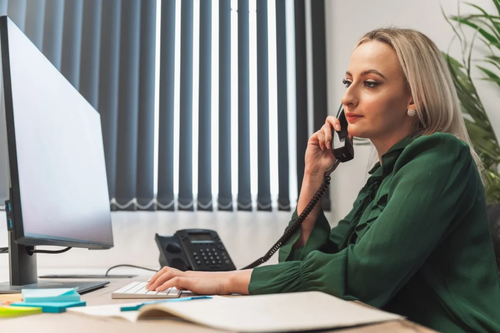 A professional woman sitting at a desk in an office, talking on the phone while working on a computer