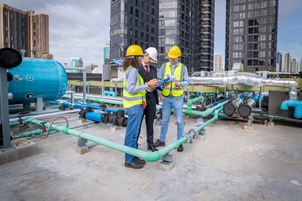 Manager and Engineers holding blueprint project under checking building cooling tower air conditioner. They check progress work chiller HVAC of large industrial building to control air system concept